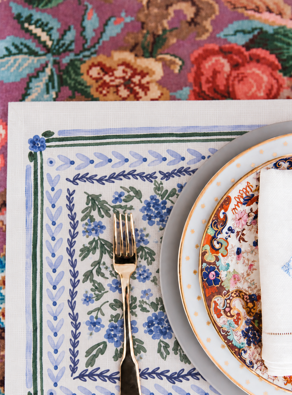 A styled tabletop setting featuring a square white linen placemat with a blue and green floral border, a gold-toned fork, and part of an ornate floral plate with a white napkin.